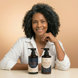 Woman with curly hair sitting behind two bottles of shampoo and conditioner on a beige background