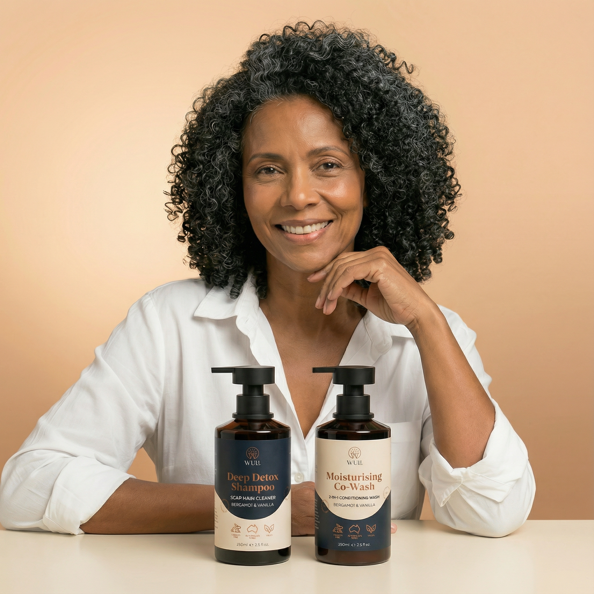 Woman with curly hair sitting behind two bottles of shampoo and conditioner on a beige background